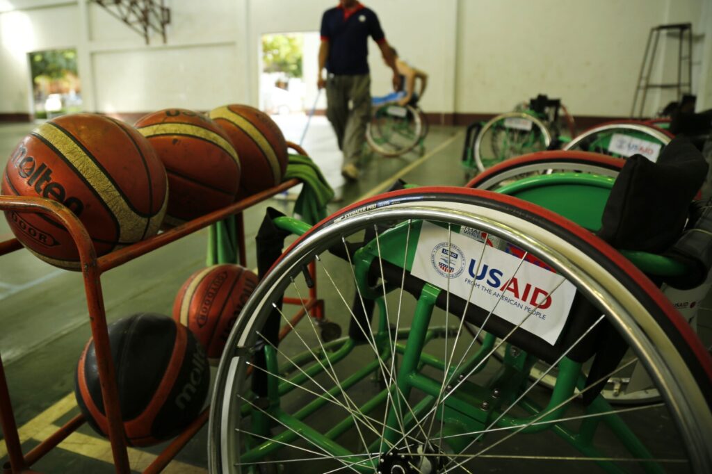 An empty USAID-branded wheelchair sits in front of a rack of basketballs on the side of a court.